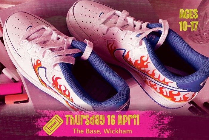 A pair of white sneakers customised with orange and red flame artwork sit on a table with paint markers nearby. A pink banner reads “Thursday 16 April – The Base, Wickham,” and text on the right says “Ages 10–17.”
