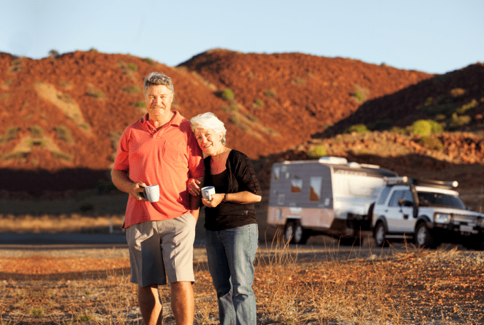 Two older adults stand outdoors holding mugs and smiling beside a camper trailer and four-wheel drive, with red rocky hills in the background under warm evening light.
